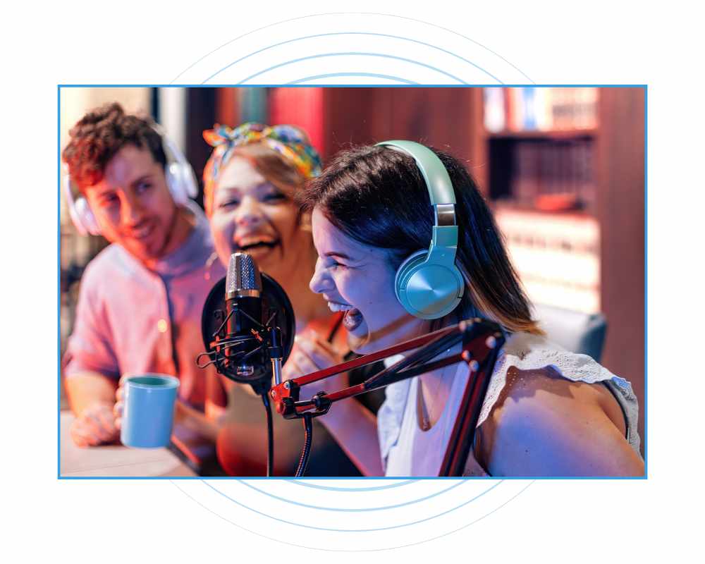 2 women and 1 man laughing in a recording studio while sitting by a microphone